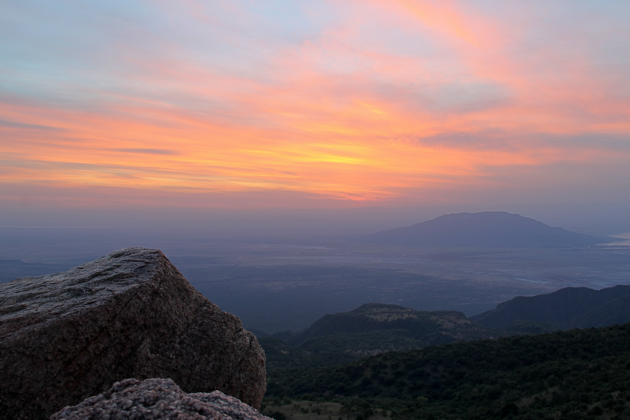 Dusk seen from a high rocky viewpoint: a large weathered granite boulder in the left foreground, forested hills falling away below, a hazy plain with scattered lights, and a long broad mountain massif on the far horizon under a pink and orange sky
