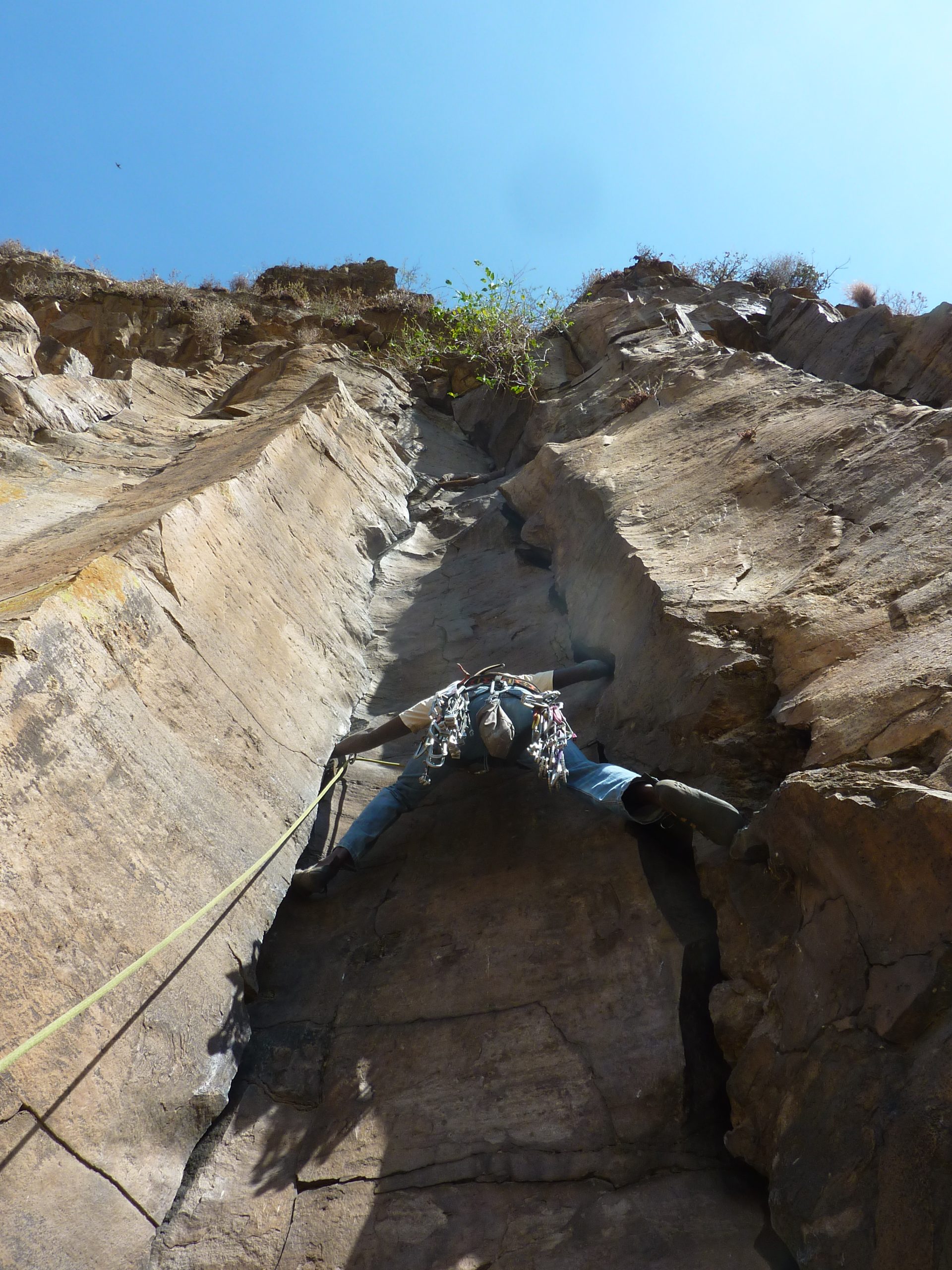 Looking straight up a sunlit gorge wall in Hell's Gate at a climber bridging a corner, one foot on each wall, a rack of cams and quickdraws slung round the waist and a yellow rope running back down out of frame. Sheer banded rock either side, blue sky and a fringe of scrub along the rim.