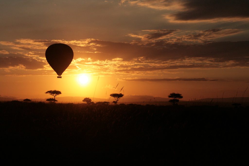 Single hot-air balloon silhouetted against a low sun at sunrise, drifting above a dark plain studded with flat-topped acacias, distant hills on the horizon