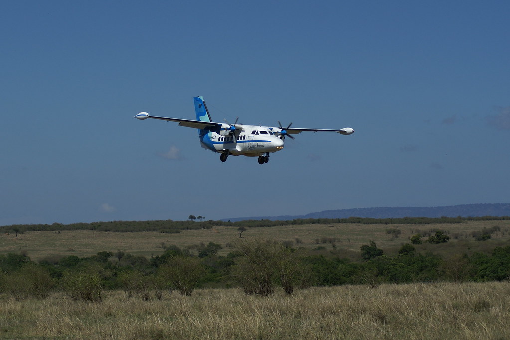 A twin-turboprop light aircraft (LET L-410 type) in white and blue, gear down on final approach, low over rolling savannah of pale grass and scattered scrub, with a long low escarpment on the horizon.