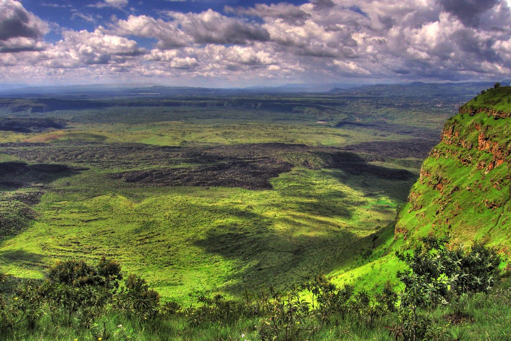 View from a grassy crater rim over a huge caldera floor of dark lava flows and bright green vegetation, with a red rock cliff on the right and cultivated highland beyond.