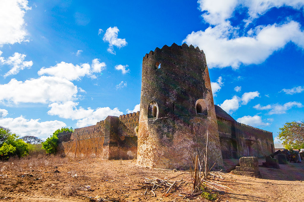 Siyu Fort on Pate Island: a tall round crenellated bastion of coral rag with narrow keyhole loopholes, joined to a long crenellated curtain wall, standing on bare red earth under a deep blue sky with cumulus cloud.