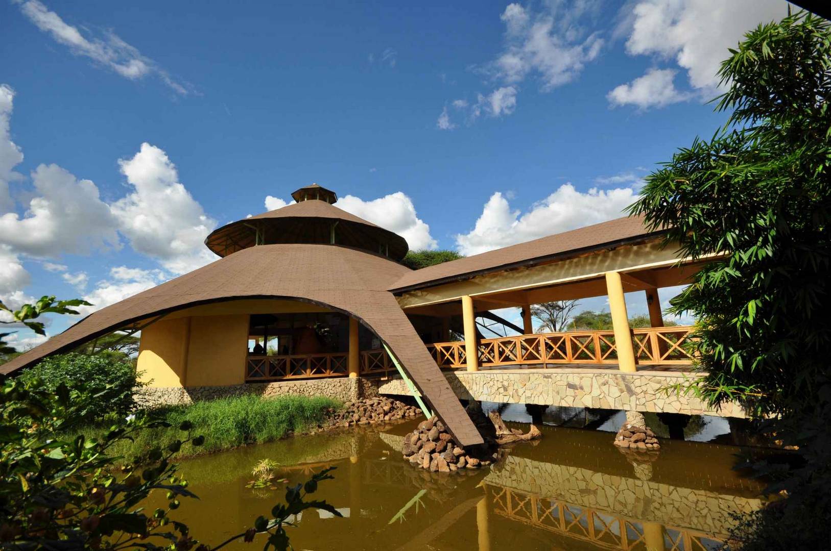 A long thatched lodge building with a dramatically curved, sweeping roof standing on stone footings above a still green pond.
